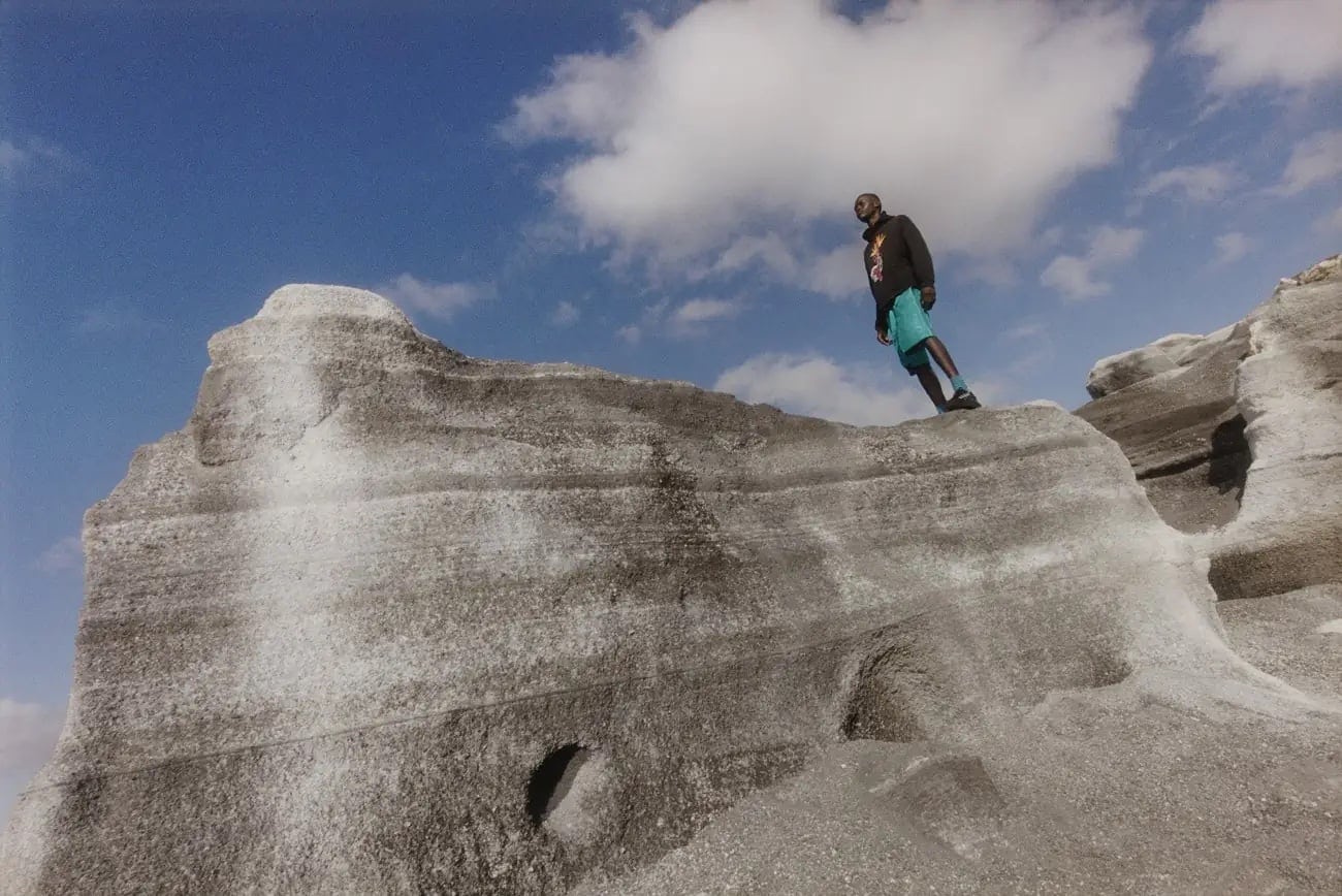 A man stands atop a large rock formation against a partly cloudy sky, wearing a dark jacket and bright turquoise shorts. 