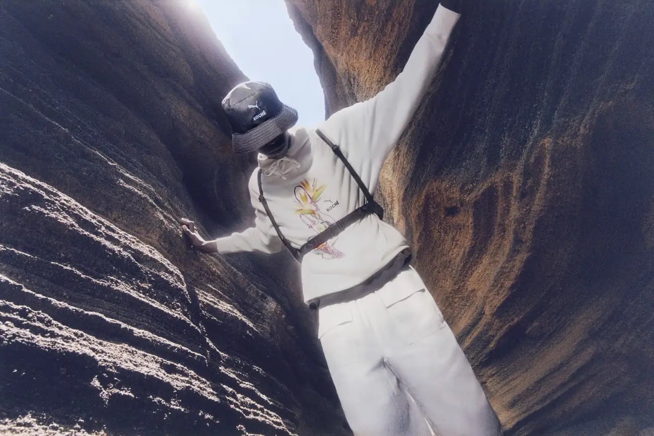 Person wearing a Puma bucket hat and sweatshirt poses in a rocky outdoor setting with a light sky in the background on a sunny day.