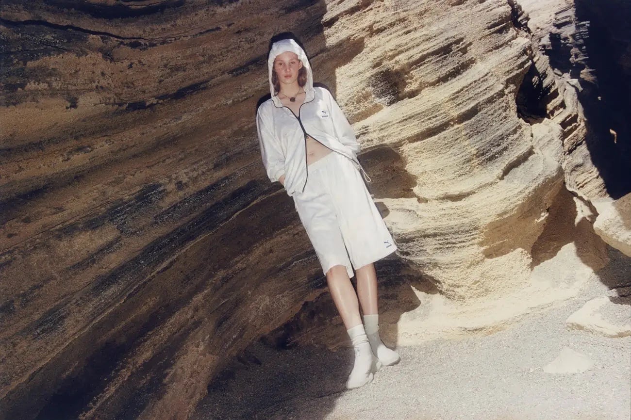 An adult woman with light skin and red hair wearing a white hooded jacket and shorts posing in front of a striated rock wall.