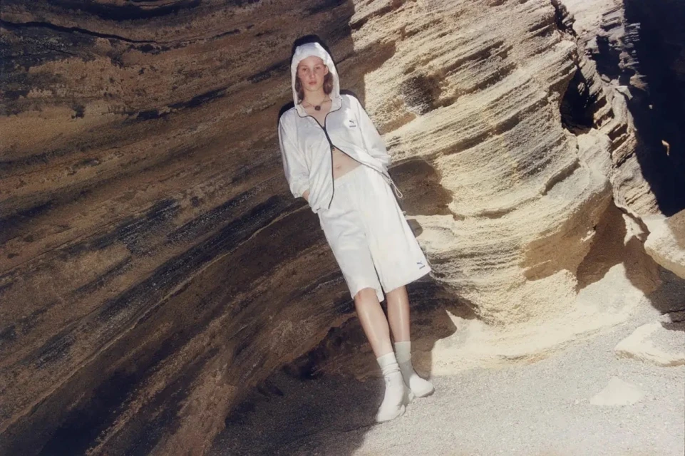 An adult woman with light skin and red hair wearing a white hooded jacket and shorts posing in front of a striated rock wall.