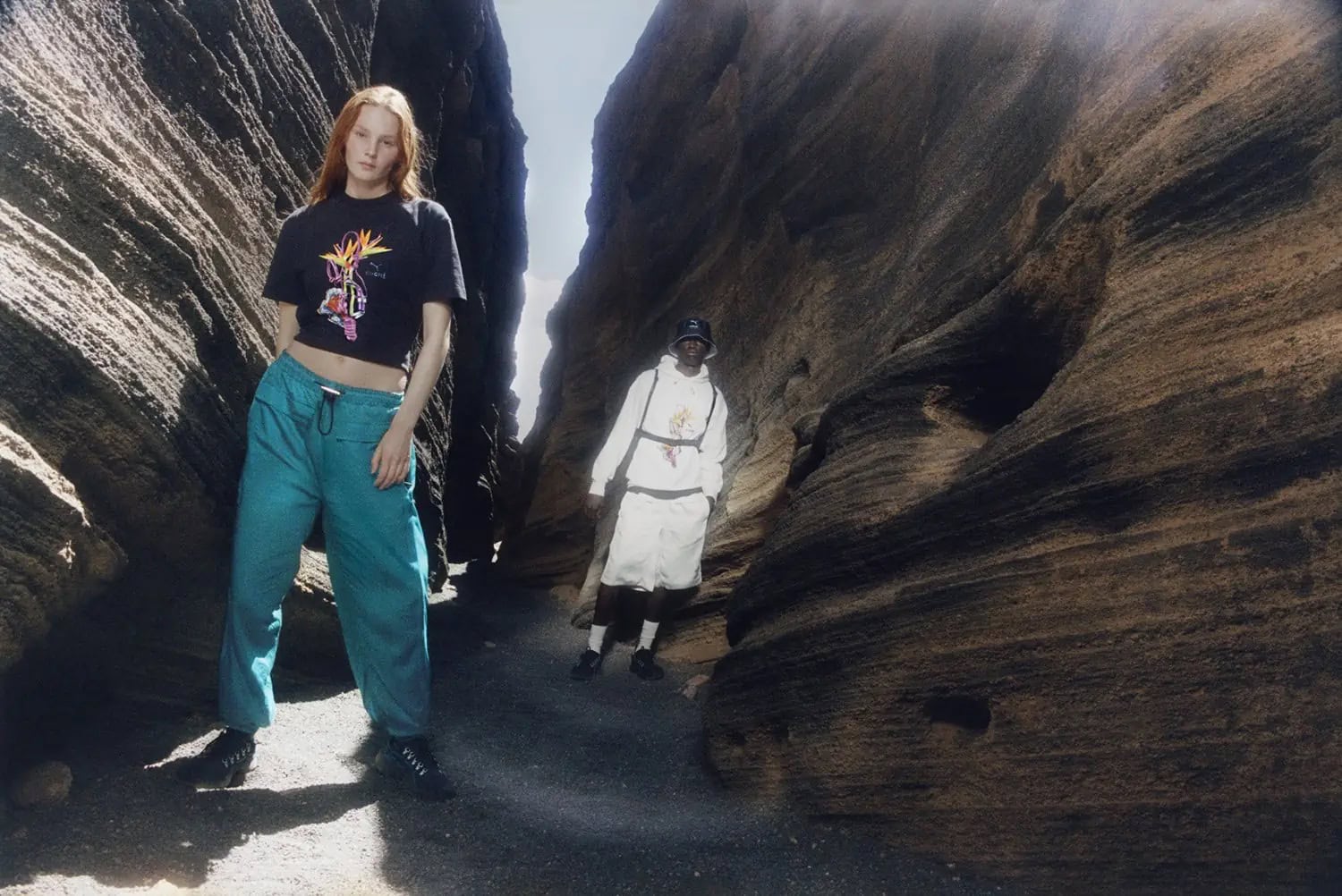 Two people pose in a natural canyon. A young woman with red hair wears a black crop top and teal pants in a full shot; fashion.