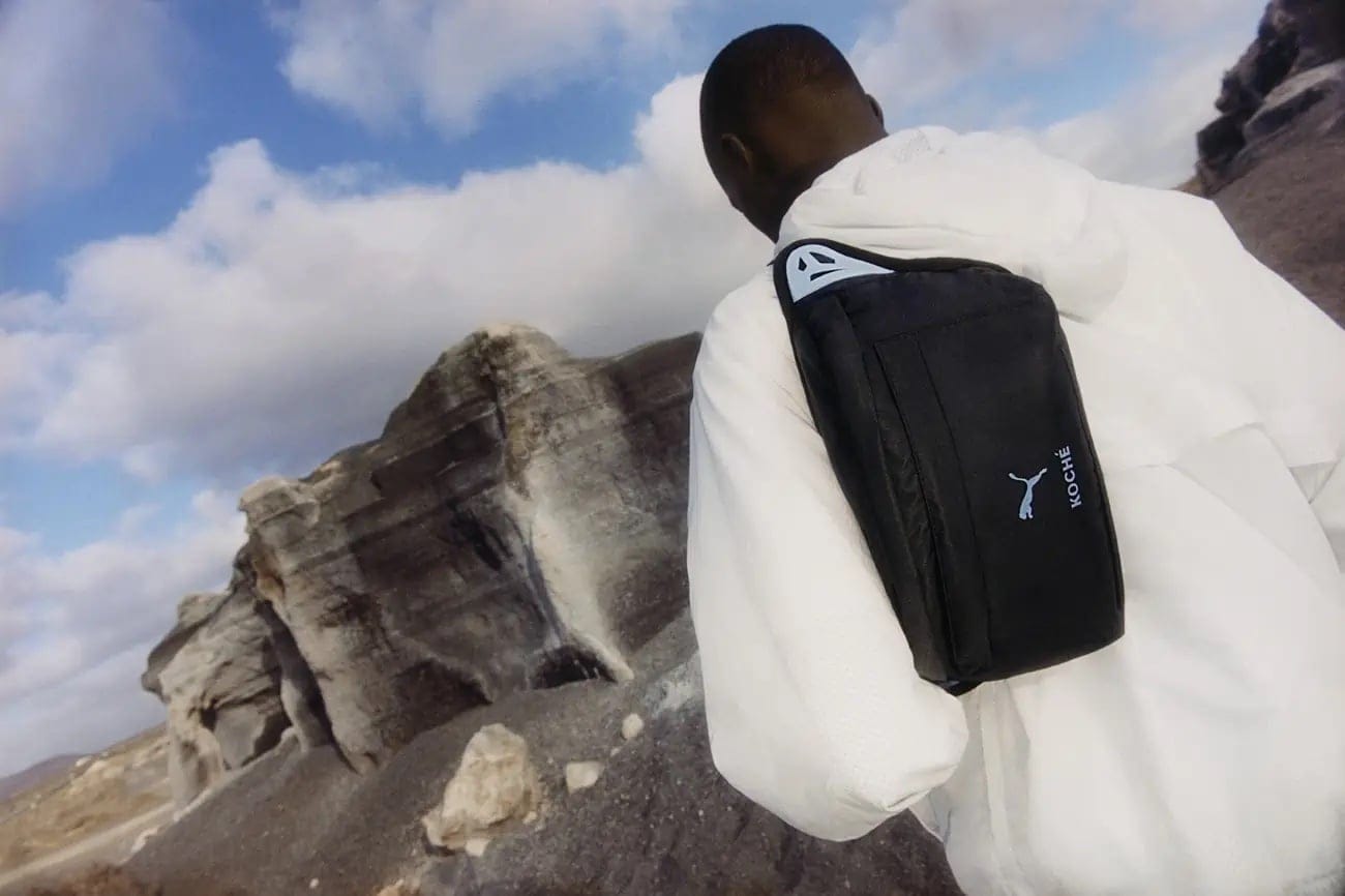 Back view of a Black male in a white hoodie outdoors with a Puma x Koché bag, set against a backdrop of striking rock formations and cloudy sky.