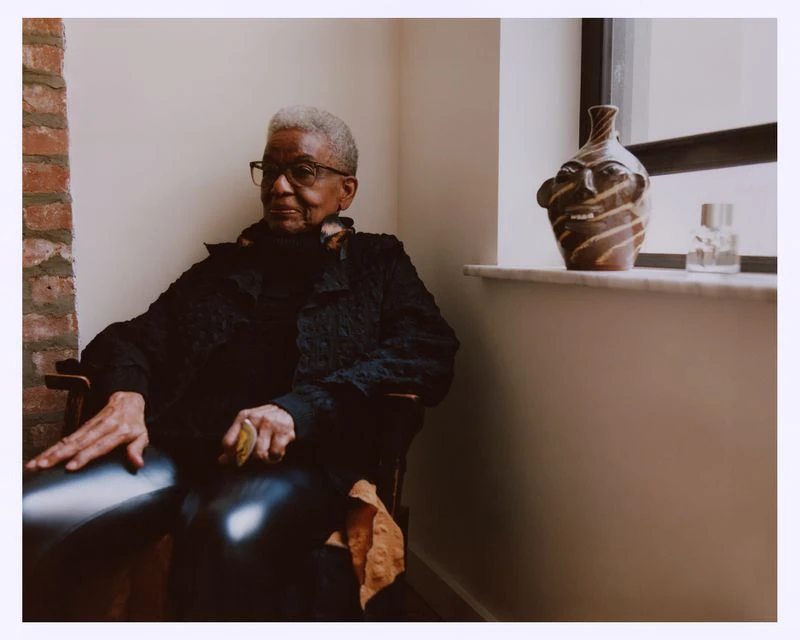 Elderly woman with short gray hair wearing glasses and a black jacket, sitting in a chair inside, next to a vase and window.