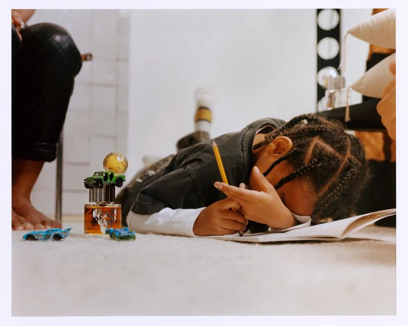 Child lies on the floor drawing with a pencil on a notepad, accompanied by a collection of toy cars and a unique perfume bottle.