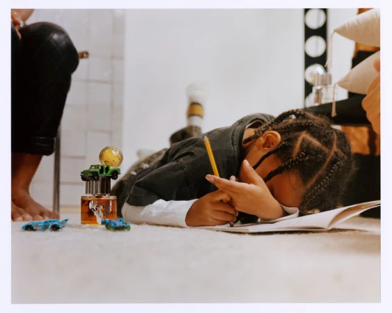 Child lies on the floor drawing with a pencil on a notepad, accompanied by a collection of toy cars and a unique perfume bottle.