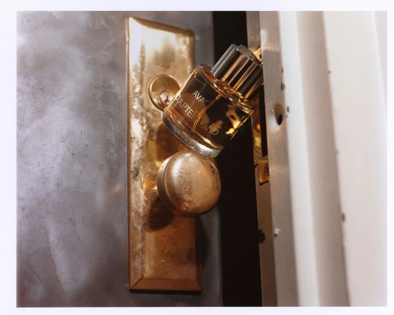 Still-life studio shot of Ava Sculpteland perfume bottle leaning on a vintage brass door handle against a gray and white door.