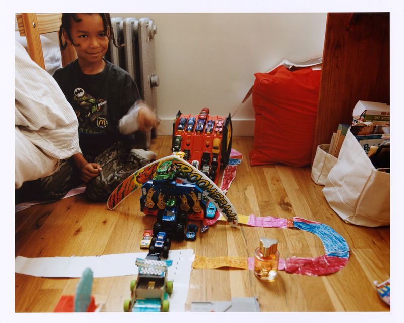 A boy plays with a toy car track on a wooden floor, surrounded by toy cars, paper decorations, and assorted household items.