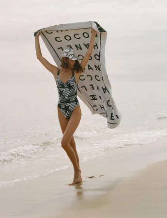Woman in swimsuit holding a Chanel towel and wearing a matching hat on a sandy beach with ocean waves in the background.