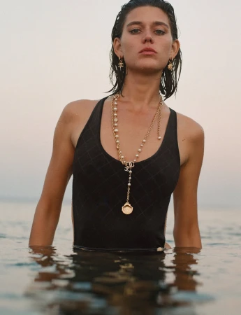 Woman in swimwear standing in water wearing Chanel jewelry, including gold chain necklace and earrings, with a soft neutral background.
