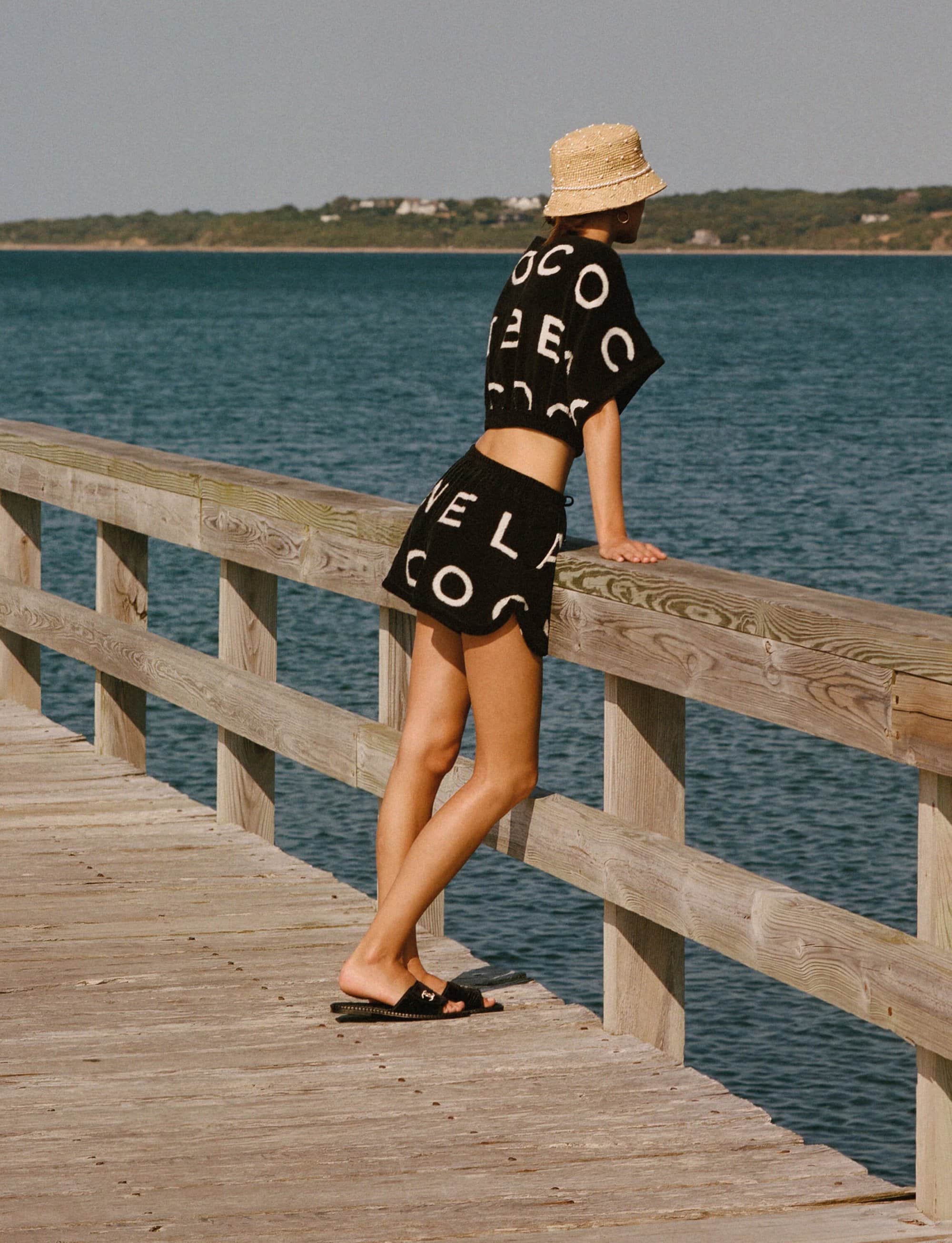 Woman in black shorts and a matching crop top leans on a wooden pier railing near the ocean on a sunny day.