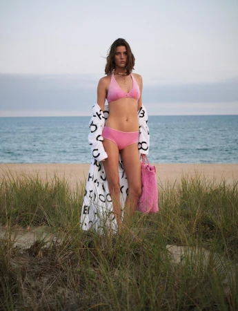 A woman in a pink bikini and patterned robe stands on a beach with green grass in the foreground and the ocean and sky in the background.