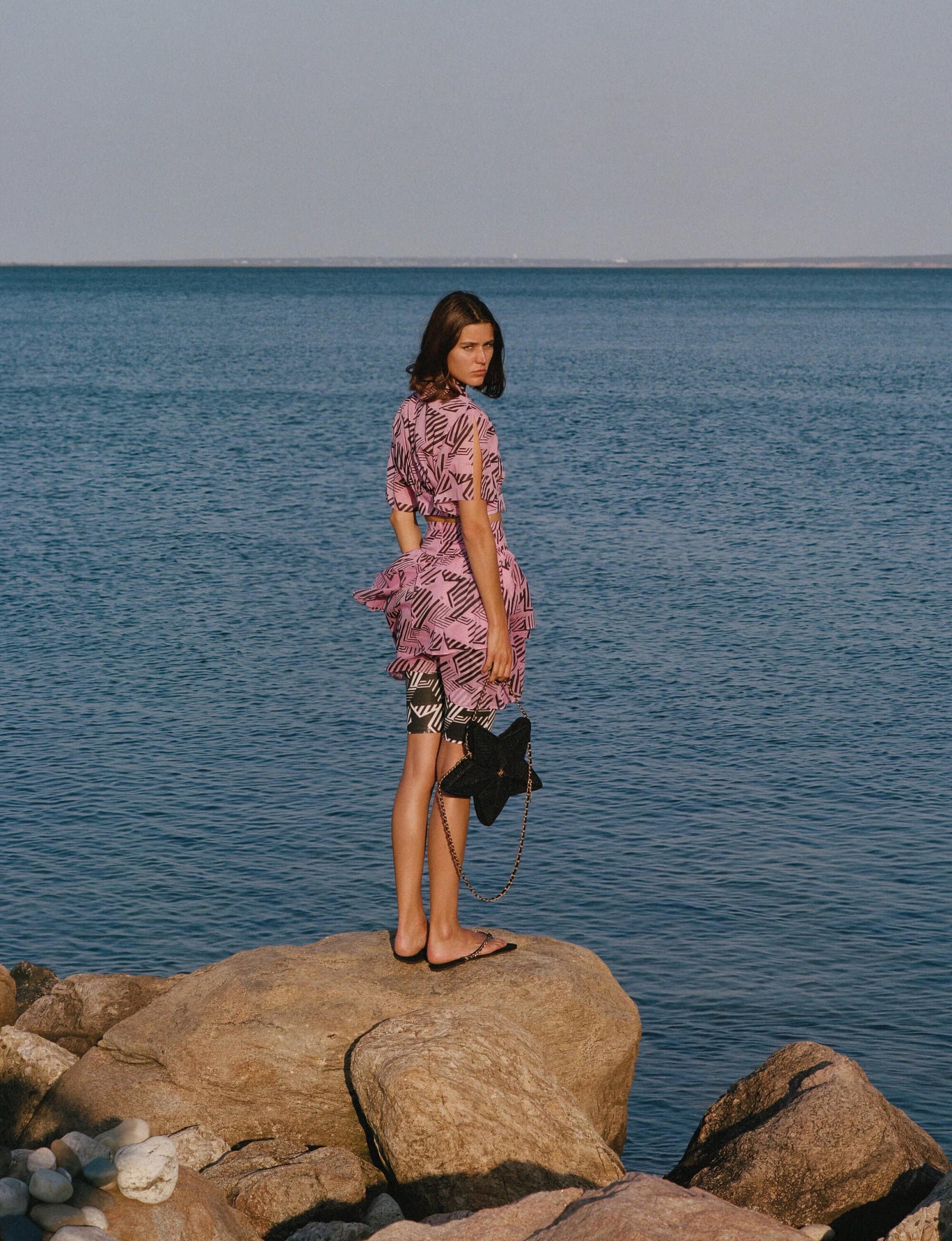 A young woman in a dress and slides stands on large rocks by the sea, looking over her shoulder, carrying a star-shaped handbag.