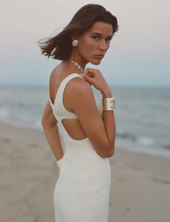 Elegant woman in white dress posing on beach with ocean backdrop, adorned with Chanel jewelry, capturing a blend of fashion and nature.