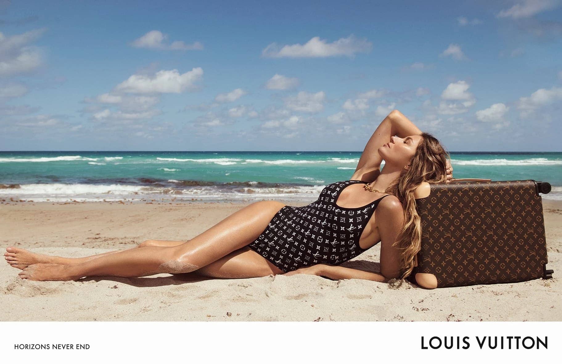 A woman in a Louis Vuitton swimsuit lounges on a beach next to a patterned suitcase with ocean and sky in the background.
