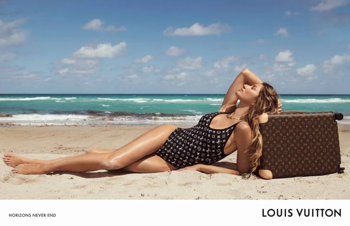 A woman in a Louis Vuitton swimsuit lounges on a beach next to a patterned suitcase with ocean and sky in the background.