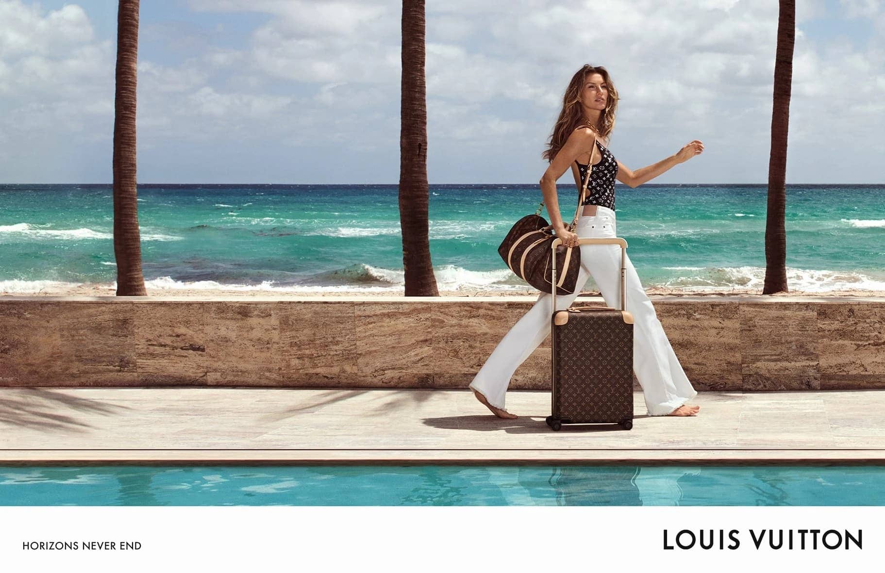 Woman walks along an oceanfront terrace with a Louis Vuitton suitcase and duffel bag, palm trees, turquoise ocean, and blue sky.