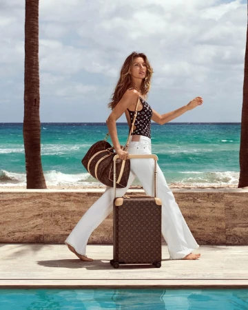 Woman walks on a beach carrying a brown patterned travel bag and rolling luggage, ocean and palm trees in the background on a sunny day.