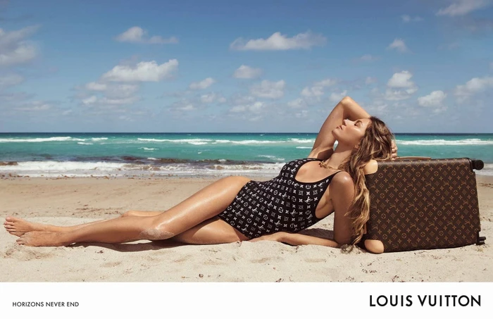 Gisele Bundchen wearing a black monogram swimsuit reclining on a sandy beach beside a brown monogram suitcase.