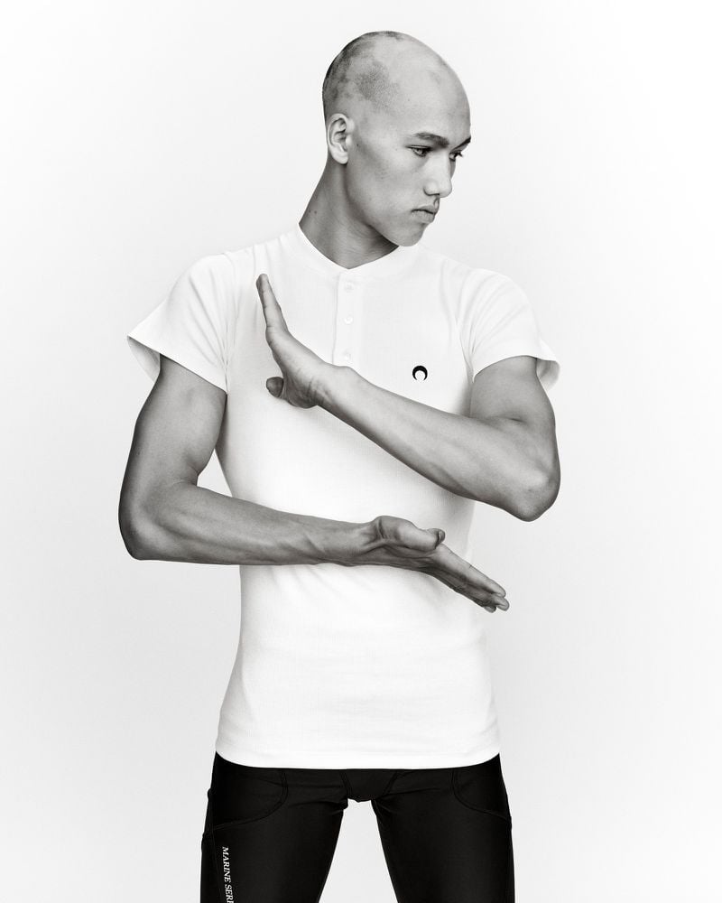 Athletic man in white shirt and black leggings poses with palms facing each other against white backdrop, showing arm muscles.