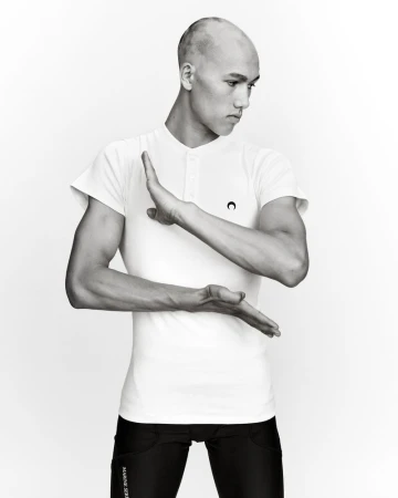 Athletic man in white shirt and black leggings poses with palms facing each other against white backdrop, showing arm muscles.