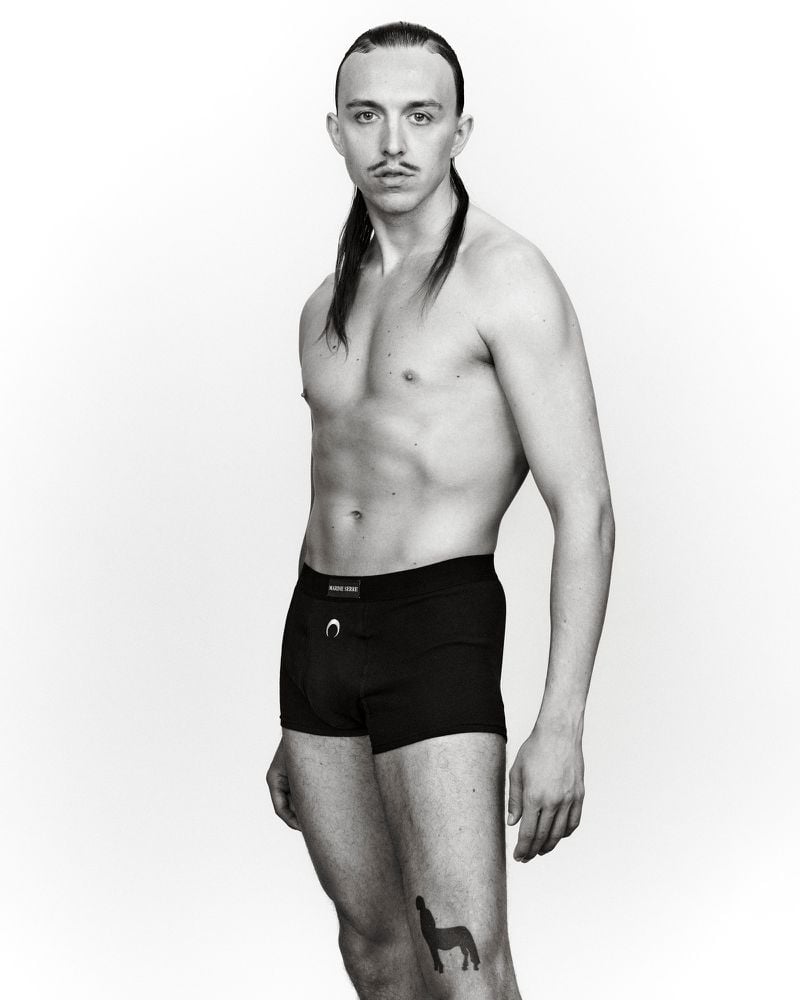 Studio shot of a slender, bare-chested adult male posing in black underwear, showcasing a centaur tattoo against a plain white background.