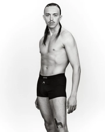 Studio shot of a slender, bare-chested adult male posing in black underwear, showcasing a centaur tattoo against a plain white background.