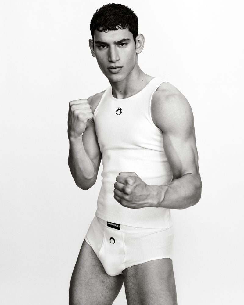 Black-and-white portrait of a young adult male in a white tank top and briefs, striking a boxing pose against a clean white backdrop.