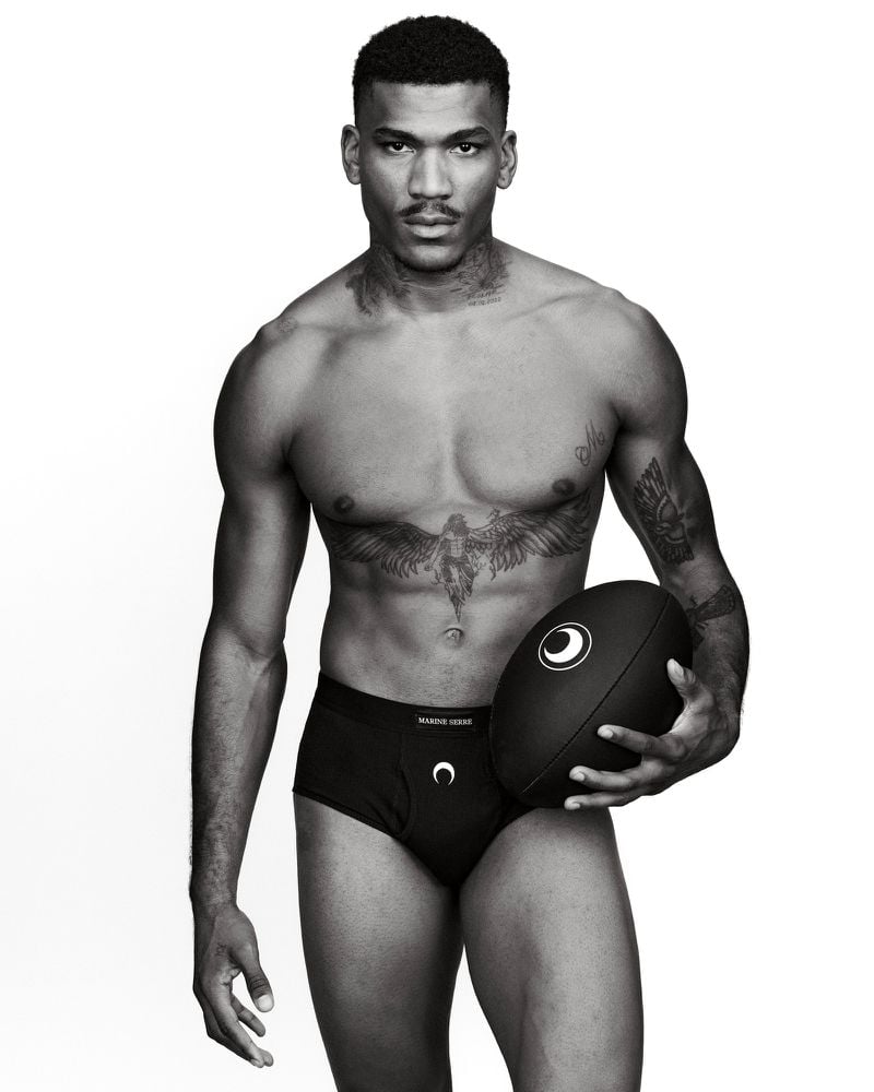 Black and white portrait of a muscular man holding a football, wearing black underwear with logos against a clean white background.