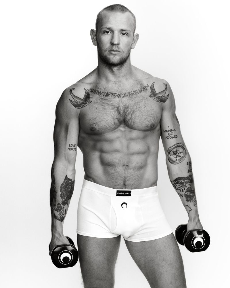 Black-and-white studio portrait of a muscular adult male posing with tattoos, wearing white underwear and holding dumbbells.