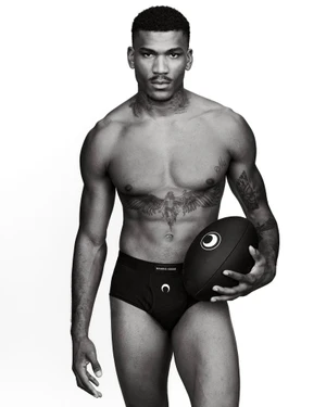 Black and white studio portrait of a shirtless Black man holding a black Marine Serre rugby ball and wearing black briefs.
