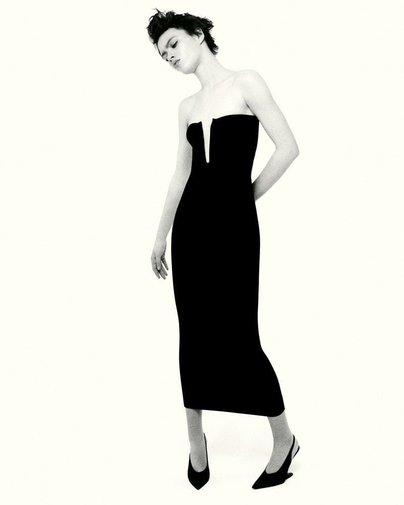 A fashionable woman in a black dress poses in a studio, showcasing an elegant design and modern silhouette against a neutral backdrop.