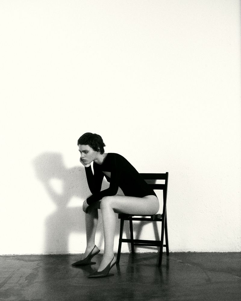 Black and white portrait of a woman in a black bodysuit and heels sitting on a chair in a minimalist photo studio.