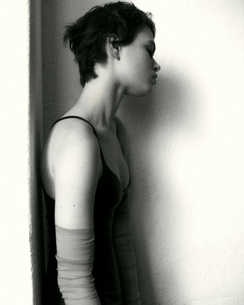 Monochrome portrait of a young adult female in a black top with short hair, leaning against a white wall, with arm coverings.