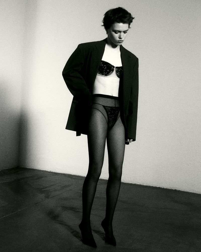 A woman in black lingerie, stockings, and an oversized jacket poses in a minimalist indoor setting with a light-colored wall.