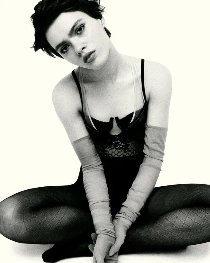Model Ayla Murray seated cross-legged wearing a black bustier bodysuit, sheer arm warmers, and diamond-pattern tights on a white backdrop.