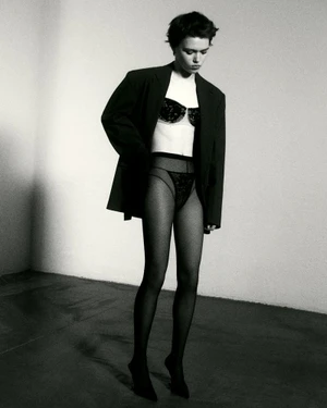 Model posing in an oversized black blazer, black lace bra, sheer dark tights, and pointed high heels in a black and white studio portrait.