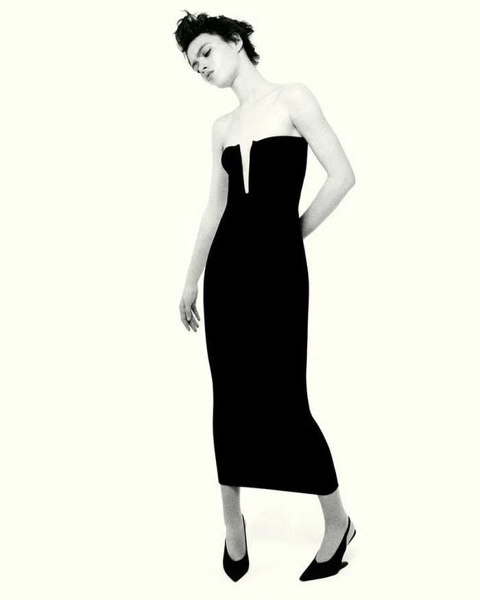 Model Ayla Murray posing in a strapless black dress with a notched neckline and pointed slingback heels against a white backdrop.