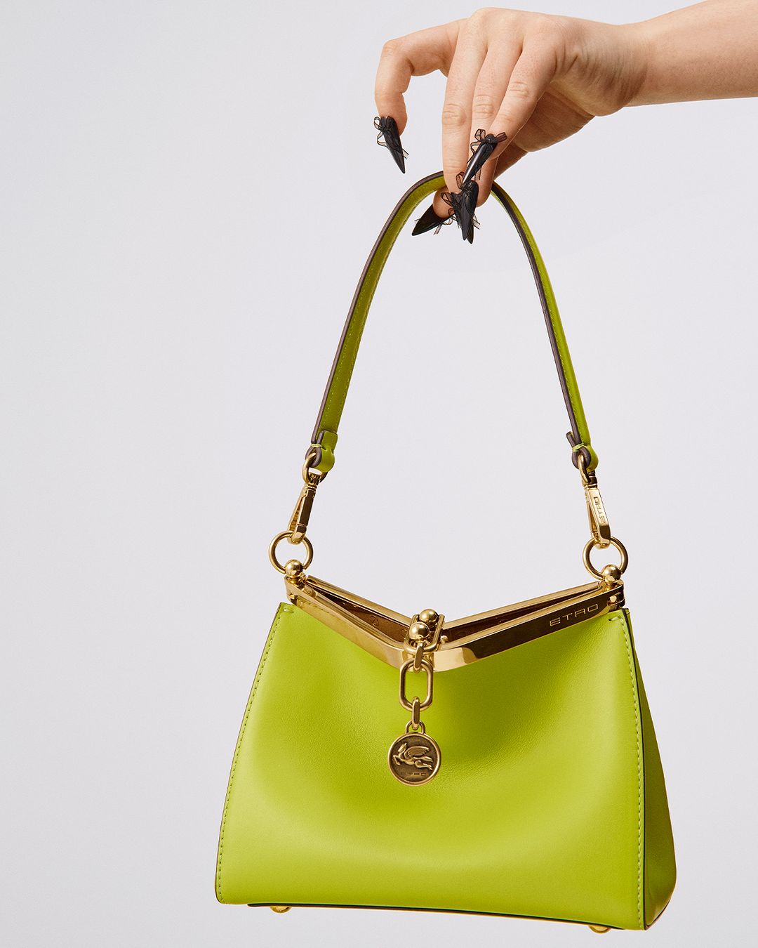 A hand with pointed black nail art holds a lime green leather Etro Mini Vela bag with gold hardware on a white background.