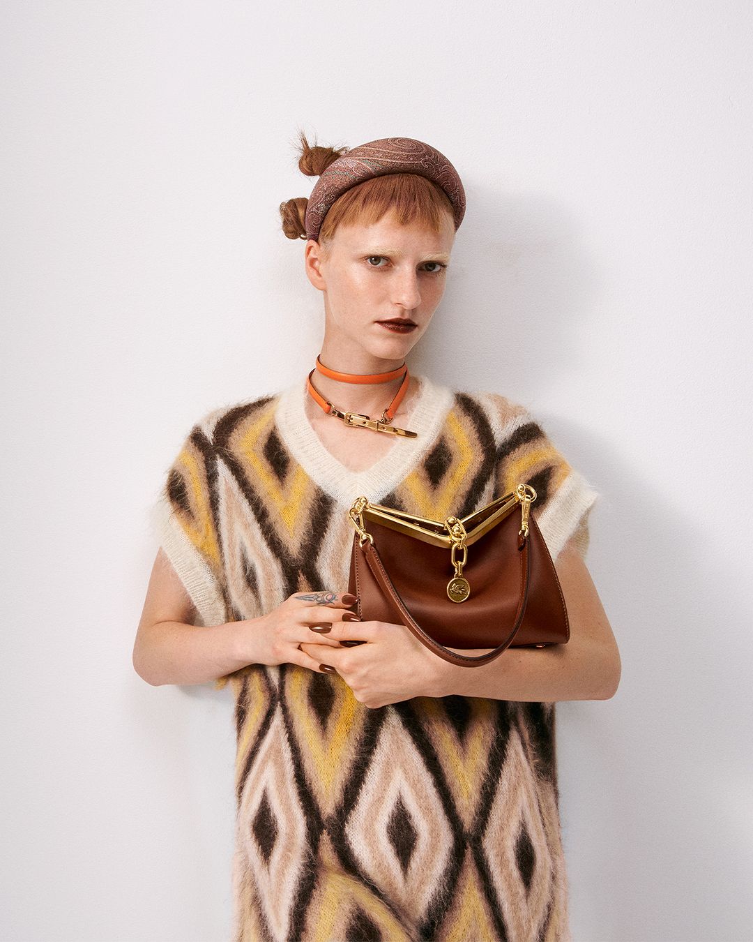 Fashionable woman presents brown leather handbag in a studio shot. Wearing patterned dress, choker and head wrap against white backdrop.