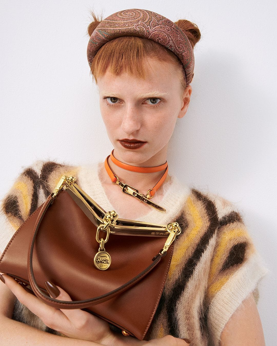 A woman with auburn hair holds a brown leather handbag with gold accents while wearing a patterned headband and colorful top.