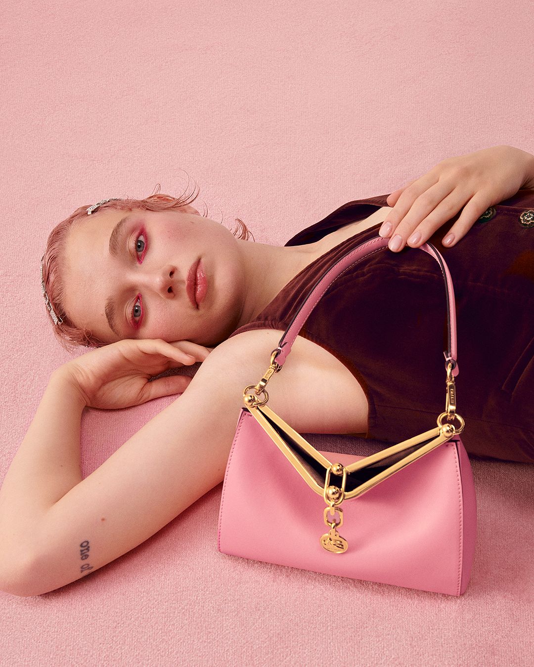A young adult woman lies on a pink textured surface next to a pink leather handbag with metal detailing in a studio setting.