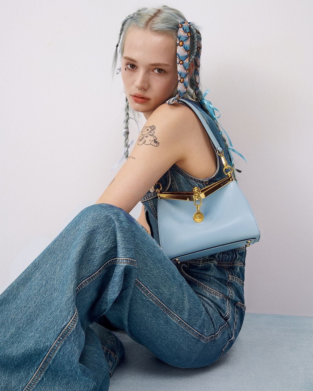 Young adult female fashion model poses with a light blue handbag, wearing a denim outfit against a pale background in a studio setting.