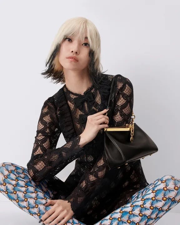 Blonde woman in lace top and patterned pants holding black purse, seated indoors against white background in portrait fashion shot.