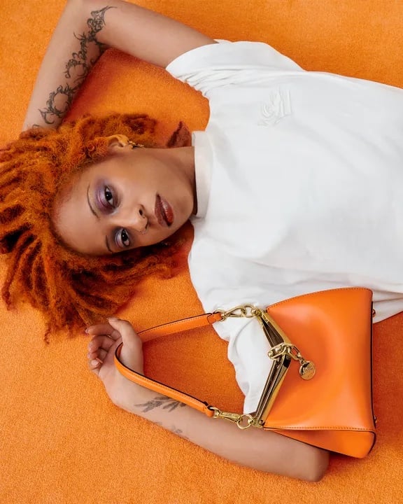 Portrait of a stylish adult woman lying on a bright orange textured background, holding a matching designer orange leather purse.