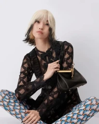 Model with blonde and black dyed hair holding a black leather handbag against a seamless white background.