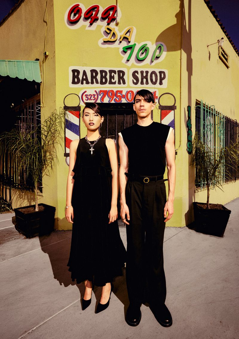 A stylish duo poses in front of a vibrant yellow barber shop, the woman in a black dress and the man in a matching sleeveless top and pants.