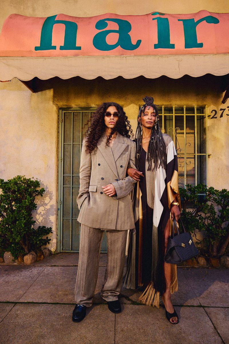 Two women stand outside a building with a 'hair' sign. One wears a gray suit, and the other wears a black and white dress with a handbag.
