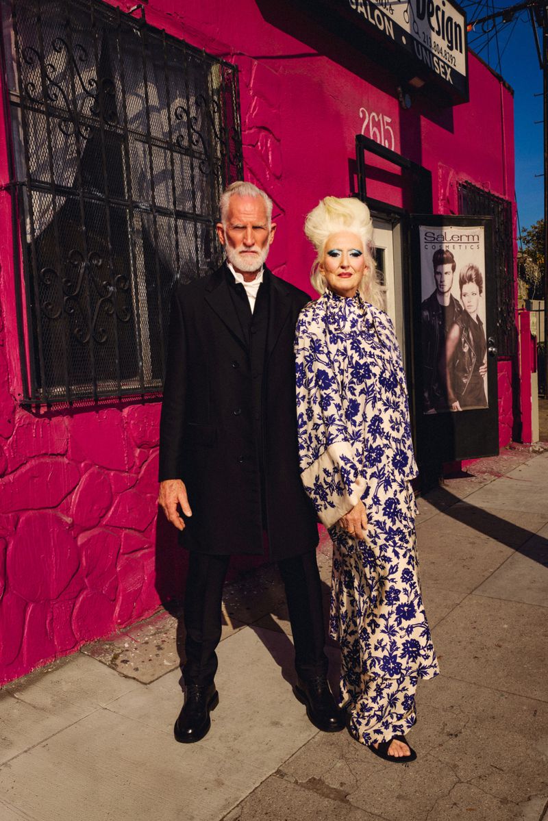 Older couple stands outside a pink building with iron-grated windows; the woman wears a floral dress, and the man wears a black suit.