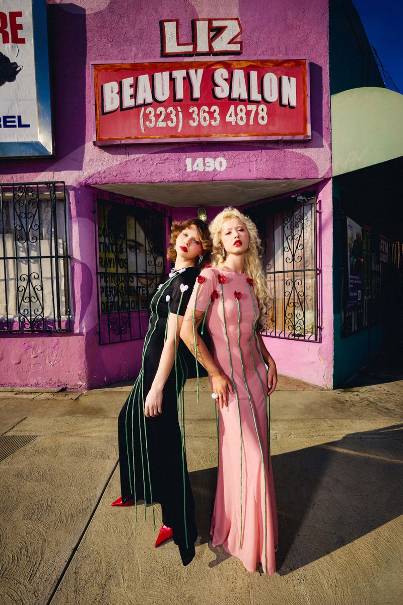 Two women pose in front of a pink building, wearing long black and pink gowns with flower details, on a sunny day outdoors.