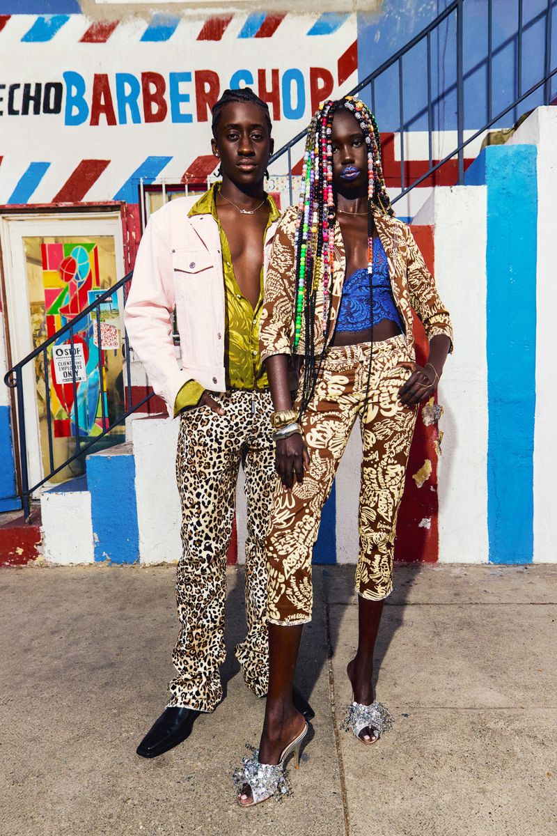 Two fashionably dressed people pose outdoors in patterned clothing, a pink jacket, and heels with a barbershop in the background.